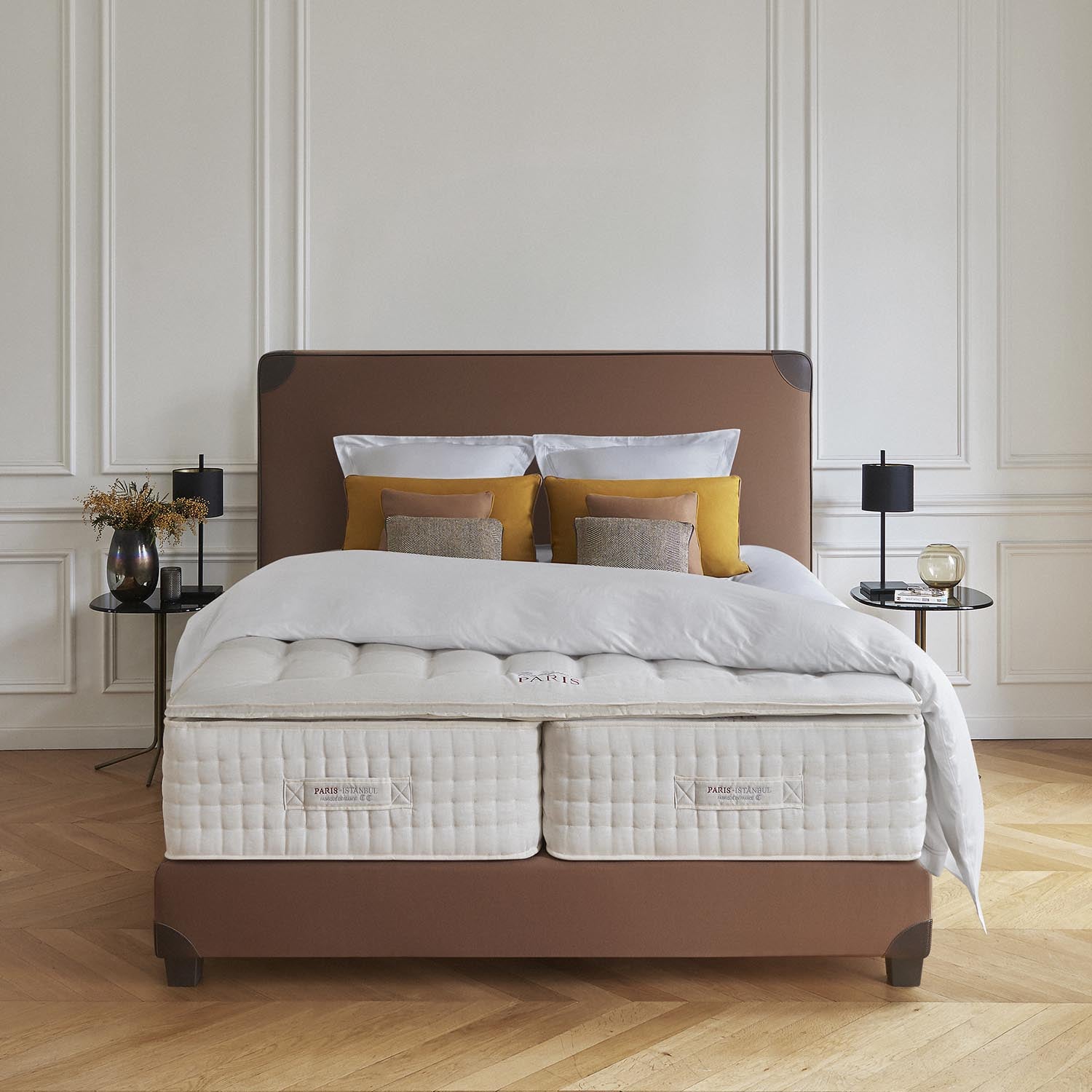 Bed with Treca Paris Istanbul mattress, brown frame, and decorative pillows in a room with wooden flooring and white walls.