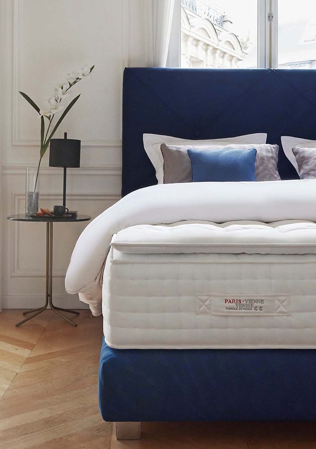 Bed with Treca Paris Vienne Venise mattress, blue headboard, and pillows in a room with a lamp and window.