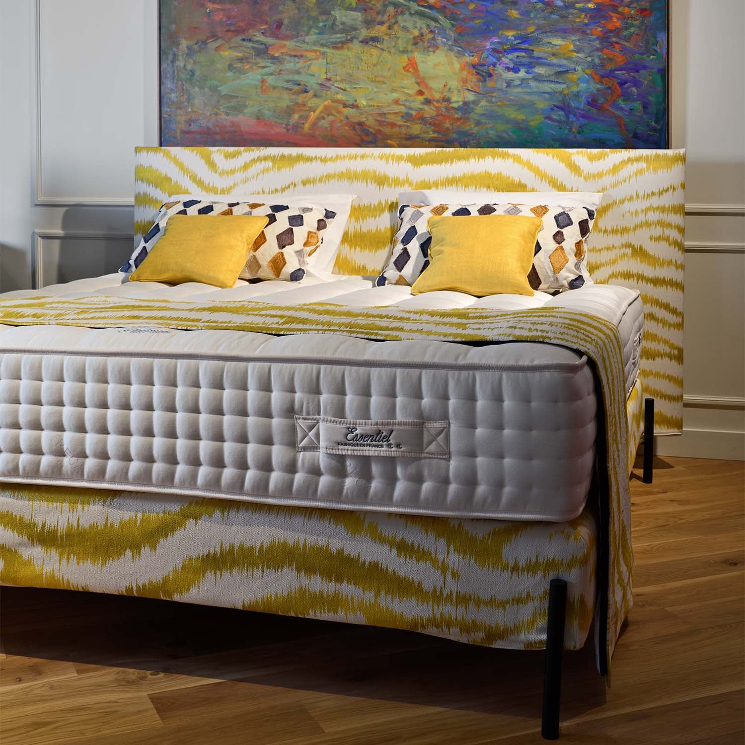 Bed with yellow and white patterned bedspread, Treca Platinum Essentiel mattress, and colorful abstract painting on the wall.